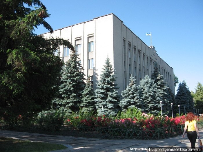 Слева от сквера — горсовет. Серое здание советского времени. Перед фасадом — голубые ели и клумбы с розами. Первомайск, Украина